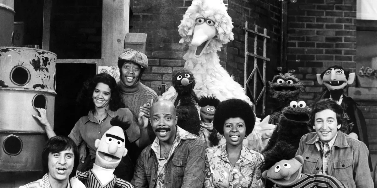 A group of Sesame Street characters and cast members, including puppets like Big Bird, Bert, Ernie, Cookie Monster, and others, posing together and smiling in a black-and-white photo.