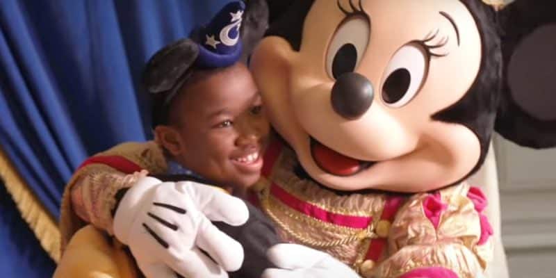A smiling child wearing a blue sorcerer hat and Mickey ears hugs a person in a Minnie Mouse costume dressed in pink and gold, with a blue curtain in the background, sharing the magic of Disney beloved by UK families.