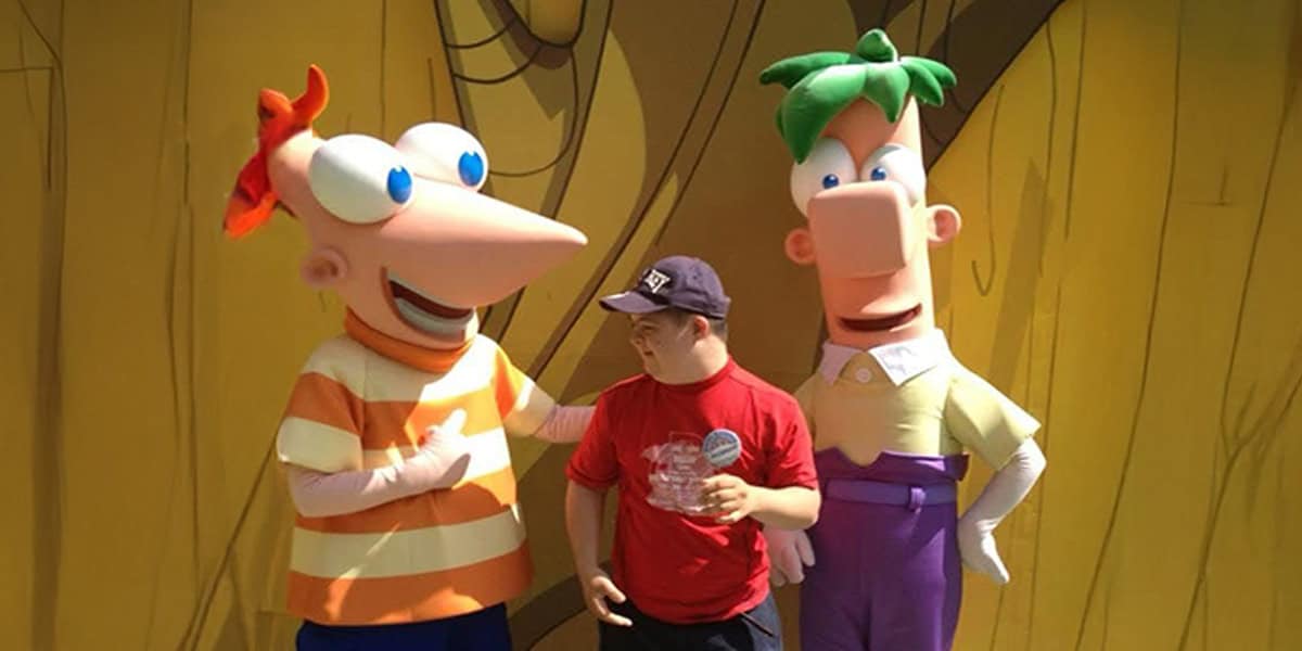 A person stands smiling between two people in Phineas and Ferb costumes at a Disney Parks meet and greet, against a yellow background with tree designs.