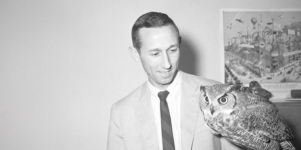 A man in a suit and tie holds a large owl on his gloved hand, looking at it. Behind him, a colorful poster with various images references Taylor Swift masters and Shamrock Capital.