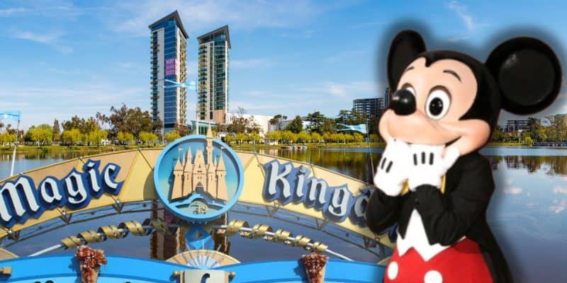 A person in a Mickey Mouse costume stands in front of the Magic Kingdom entrance sign, with a lake, trees, and tall buildings under a clear blue sky—a perfect scene to highlight Disney World vacation fees on a new website.