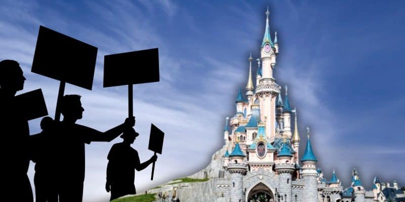 Silhouetted protesters holding signs stand in front of a fairytale castle with blue rooftops under a blue sky with wispy clouds inside of Disneyland Paris as taxi strike takes place.