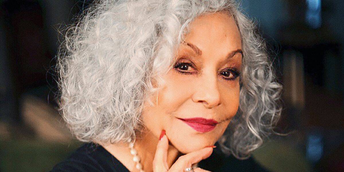 Renee Victor with curly gray hair and red lipstick smiles gently, resting her chin on her hand. Reminiscent of Abuelita from Disney Pixar Coco, she wears a black top, a pearl necklace, and has red nail polish. The background is softly blurred.