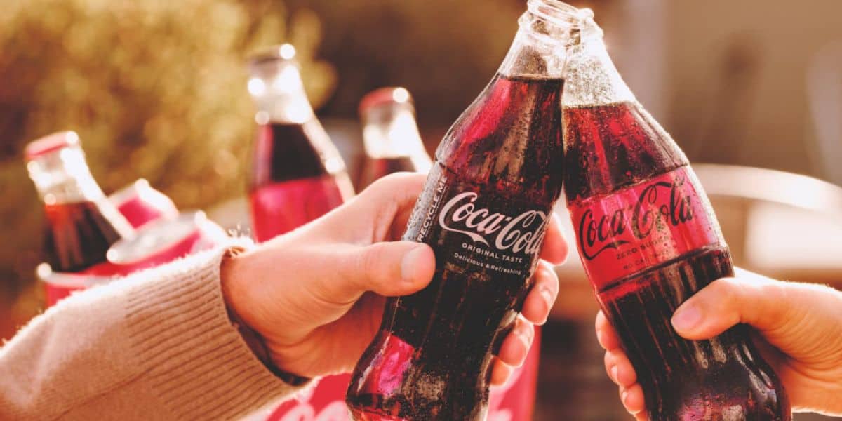 Two hands clink glass Coca-Cola bottles together in a toast, with more Coke bottles and a blurred outdoor background visible—a refreshing moment after a fun day exploring Disney Park Prices. Condensation on the bottles shows they're ice-cold.