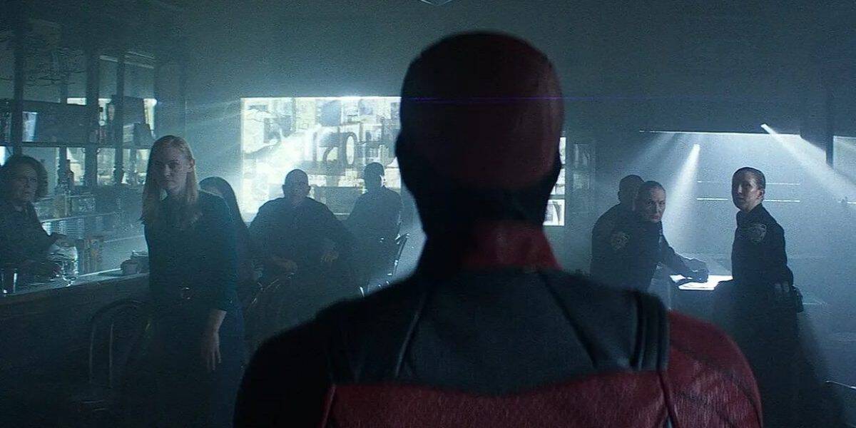 A man in a red superhero suit stands with his back to the camera, facing a group of people and police officers inside a dimly lit diner. Several people turn to look at him, appearing surprised or cautious.