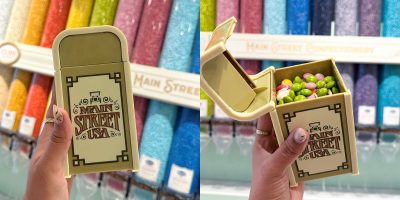 A hand holds a Disney World Trash Can-themed container shaped like a tiny Main Street USA trash can, filled with colorful jelly beans, with rows of candy dispensers visible in the background.