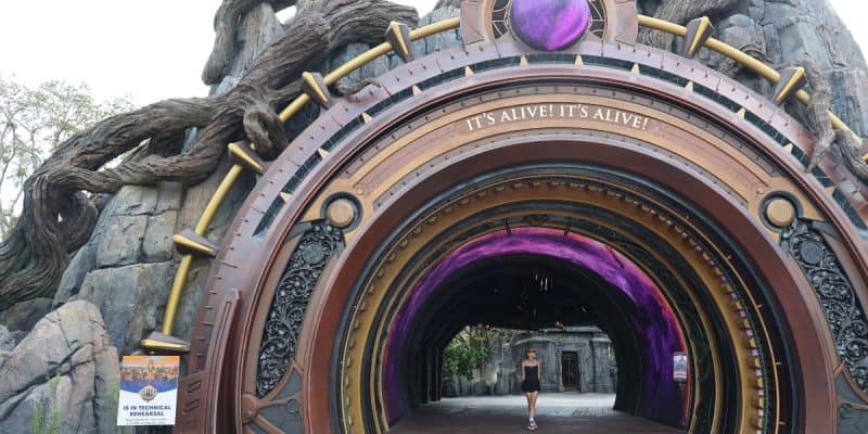 A person stands at the entrance of an elaborate, circular, steampunk-themed archway at Epic Universe, with "IT'S ALIVE! IT'S ALIVE!" above. The tunnel glows with purple light, surrounded by rocky structures and twisted tree roots.