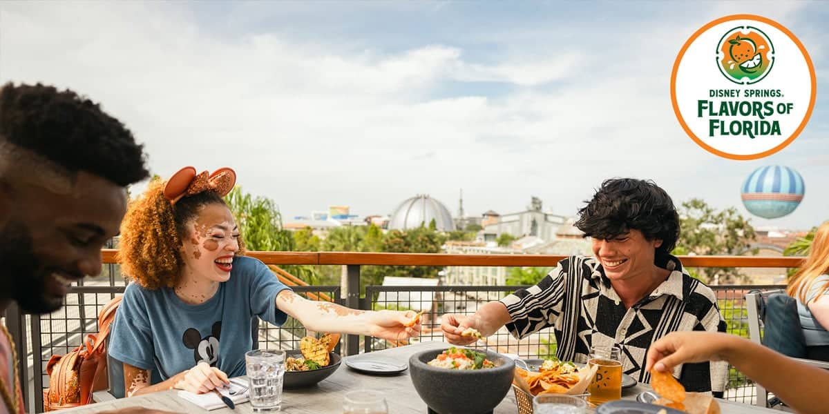 A group of friends enjoy food and laughter at an outdoor restaurant with a view during Disney Springs Flavors of Florida. The table is filled with dishes and drinks under a sunny sky, celebrating the tastes of Summer 2025.