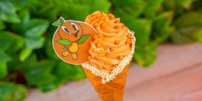 An orange ice cream cone topped with orange swirled frosting, candy pieces, white sprinkles, and a decorative edible disc featuring a smiling cartoon orange character celebrates Flavors of Florida at Disney Springs, perfect for Summer 2025.
