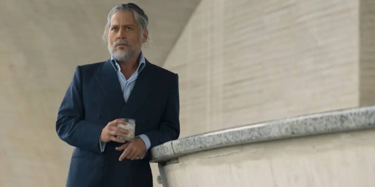 A man with gray hair and a beard, dressed in a dark suit, stands by a curved stone railing holding a drink, looking thoughtful in a modern, minimalist setting reminiscent of filming scenes with Johnny Depp from the Pirates franchise.