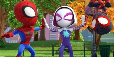Three cartoon superheroes, inspired by Spider-Man, are in a park. Two face each other giving high-fives, while a third hangs upside down from a streetlamp. Bright trees and buildings complete this Disney+ animated slate scene.