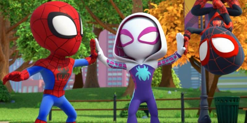Three cartoon superheroes, inspired by Spider-Man, are in a park. Two face each other giving high-fives, while a third hangs upside down from a streetlamp. Bright trees and buildings complete this Disney+ animated slate scene.