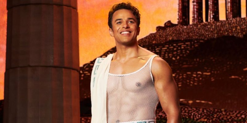 A smiling man stands confidently in front of a backdrop resembling ancient ruins, evoking scenes from Hercules Musical. He wears a white mesh tank top and a sash draped over one shoulder, while the warm lighting adds a dramatic effect.