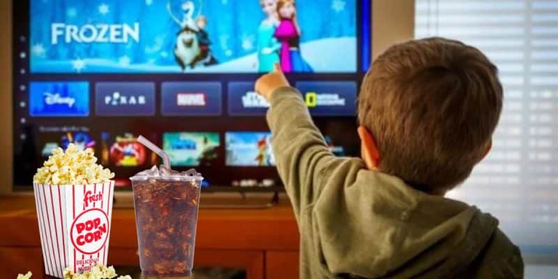 A child points at a TV screen displaying the movie "Frozen" and Disney+ alongside various streaming apps. In the foreground are a tub of popcorn and a cup of soda, reminiscent of treats from concession stands.