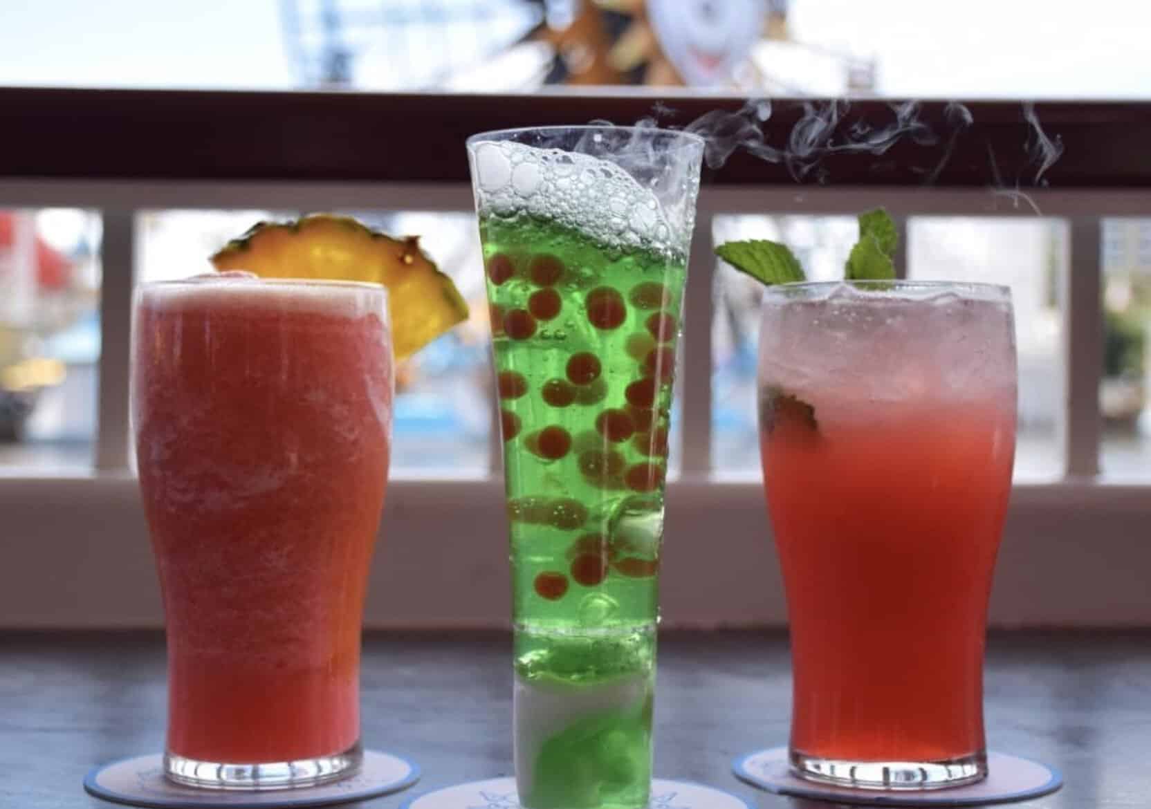 Three colorful drinks at Lamplight Lounge: a pink drink with pineapple, a tall green bubbly drink with red spheres and a smoky effect, plus another pink drink with mint. Secure your Lamplight Lounge reservations to enjoy these unique creations.