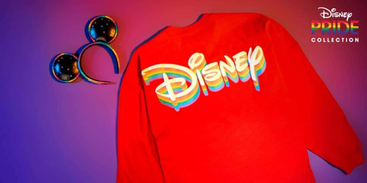 A red shirt with a rainbow-colored Disney logo and Mickey Mouse ears, set against a colorful background. The Disney Pride Collection logo appears in the top right, celebrating Disney LGBTQ+ pride and inclusivity.
