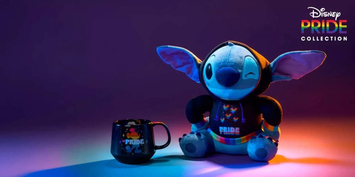 A plush Stitch toy in a rainbow "Pride" outfit sits beside a matching black mug under colorful lighting, showcasing joyful Disney Pride merch. The Disney Pride Collection logo appears in the top right corner.