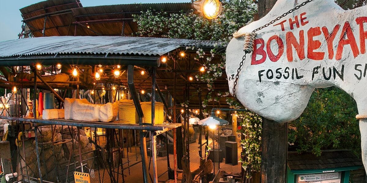 At the Boneyard Animal Kingdom, a large stone sign shaped like a dinosaur bone reads "The Boneyard Fossil Fun Site Est. 1947," welcoming guests to a fossil dig play area decorated with lights and excavation tools.