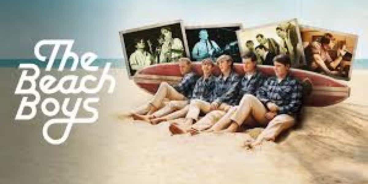 Five men in matching plaid shirts and khaki pants sit on a surfboard on the beach. "The Beach Boys" is written on the left, with vintage photos of the band playing music shown above them.