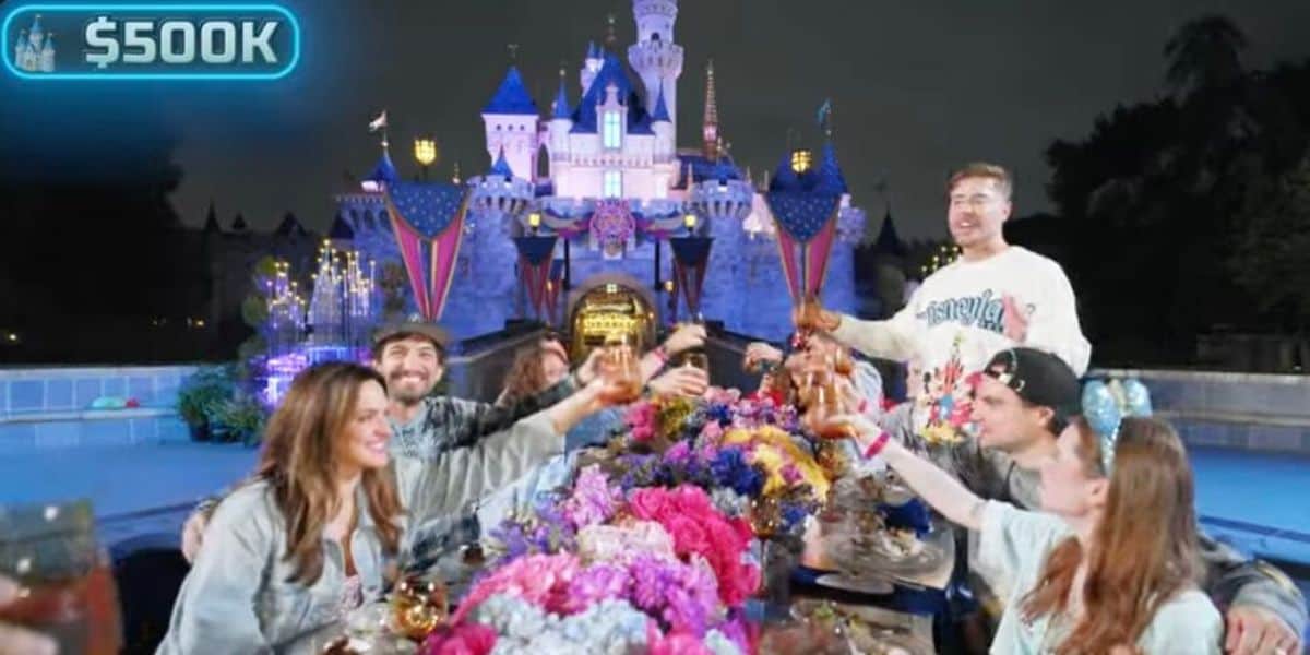A group of people cheer and raise their glasses around a colorful, flower-filled table in front of a brightly lit castle at night. A "$500K" label appears in the top left corner.