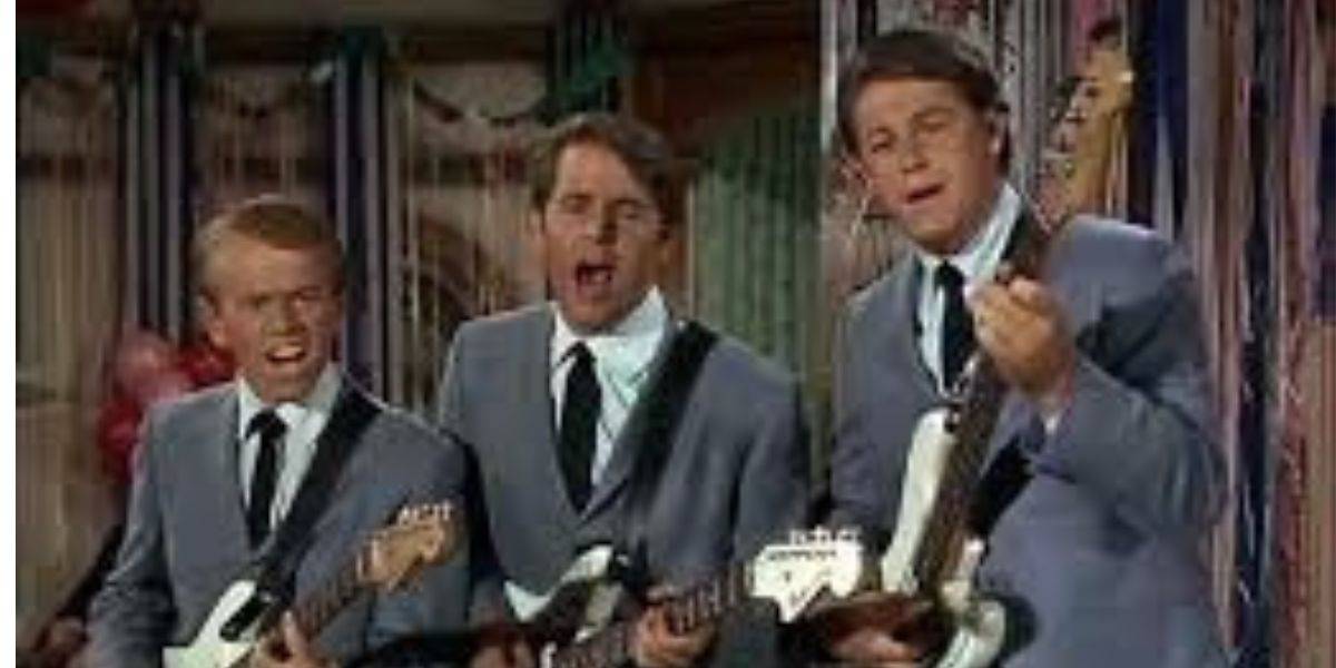 Three men in matching gray suits and black ties play electric guitars and sing into a microphone, performing together on a decorated stage.