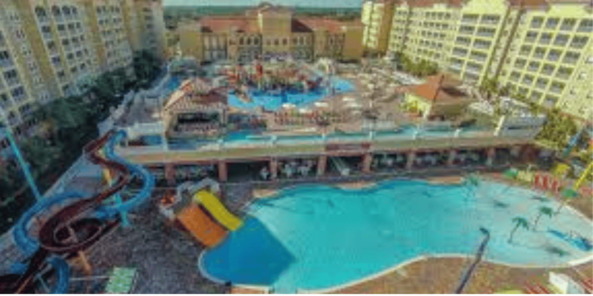 Aerial view of a large resort with several pools, water slides, lounge chairs, and tall yellow hotel buildings surrounding the water park area.