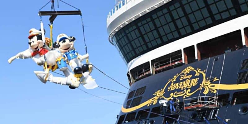 Two large Mickey and Minnie Mouse mascots dressed as ship captains are suspended by a crane beside the Disney Adventure cruise ship, with crew members working below.