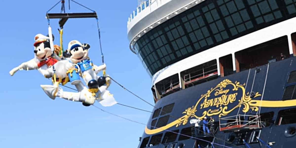 Two large Mickey and Minnie Mouse mascots dressed as ship captains are suspended by a crane beside the Disney Adventure cruise ship, with crew members working below.