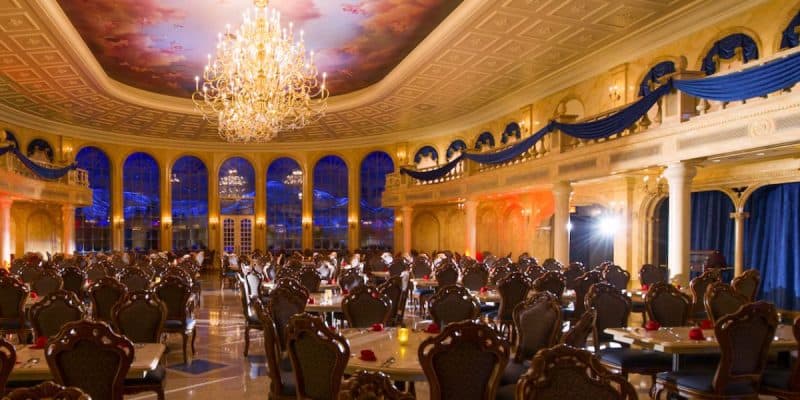 A grand ballroom with ornate chandeliers, elegant arched windows, luxurious blue drapes, and dozens of intricately carved chairs and tables set for dining under a painted ceiling—reminiscent of a Disney World malfunction frozen in time.