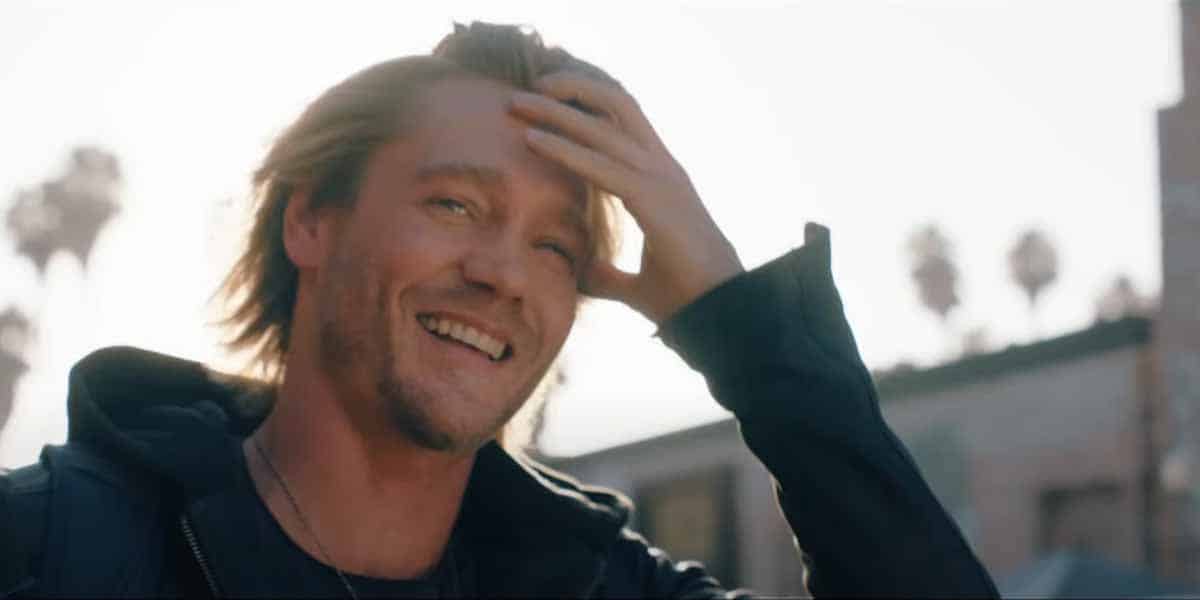 A smiling man with shoulder-length blond hair runs his hand through his hair while standing outdoors on a sunny day, as if he's just seen the latest Freakier Friday 2025 trailer; palm trees and buildings blur in the background.