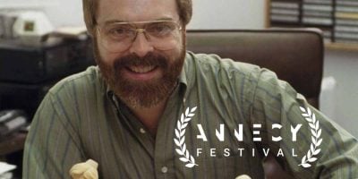 A bearded man with glasses smiles while sitting at a desk with sculpted figures. The Annecy Festival logo with laurel branches and the Annecy Walk of Fame are overlaid in the foreground.