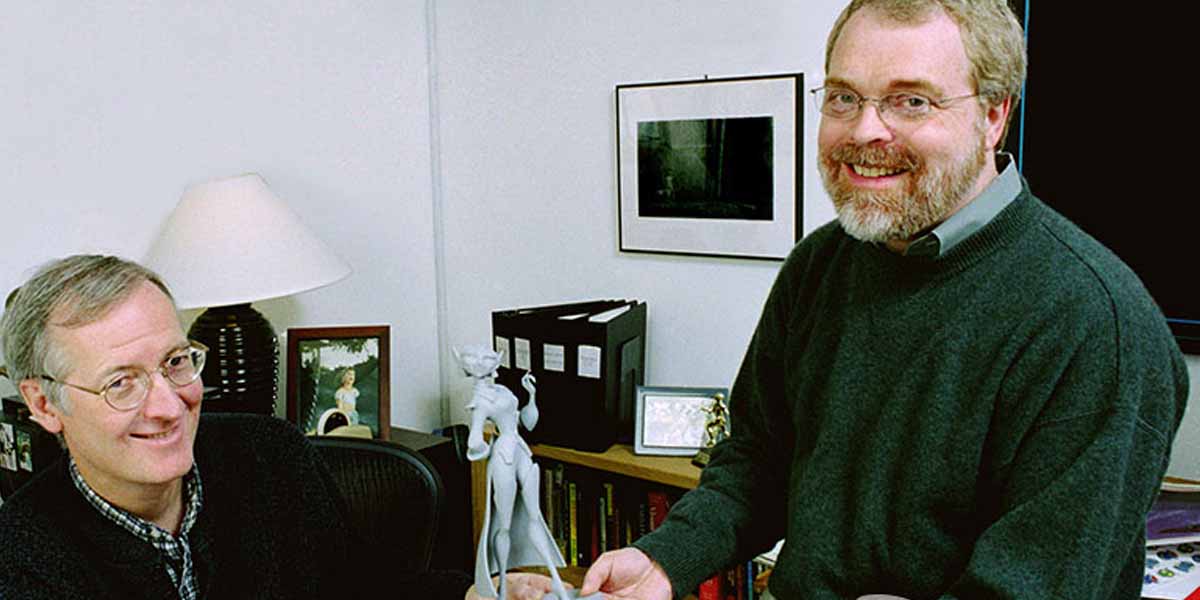 Two men in sweaters smile in an office. One is seated at a desk, while the other stands holding a figurine. Books, framed photos, and office items are visible—perhaps discussing Ron Clements' upcoming films or the Annecy Walk of Fame honor.