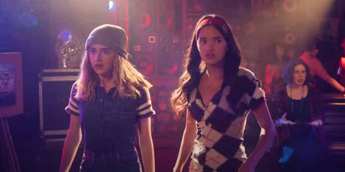 Two teenage girls stand side by side in a dimly lit room with colorful lights and speakers behind them, looking ahead with serious, concerned expressions—capturing the tense mood teased in the Freakier Friday 2025 trailer. Another person sits in the background.