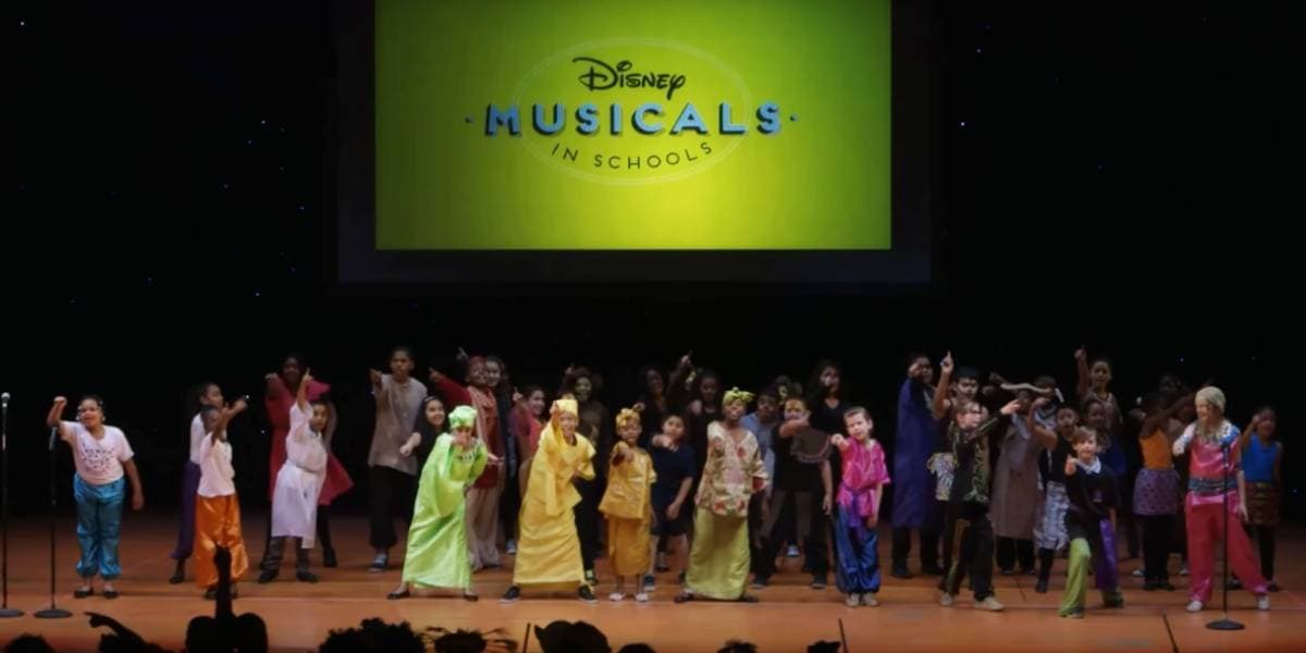 A large group of children in colorful costumes perform on stage under the "Disney Musicals in Schools" logo, singing and gesturing energetically against a dark backdrop.