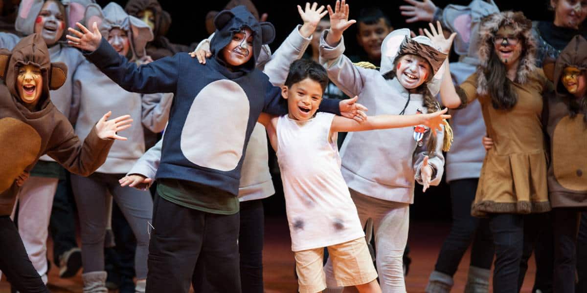 Children dressed in animal costumes smile and pose on stage during a school play inspired by Disney Musicals in Schools, with one child in the center wearing a plain white outfit and spreading his arms wide.