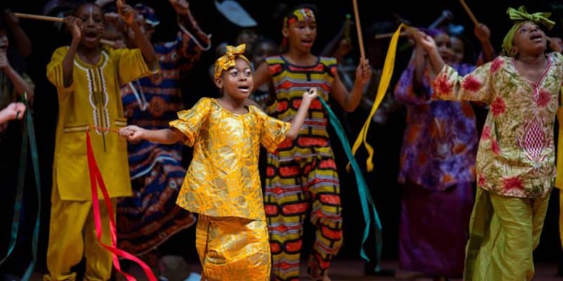 A group of children in colorful traditional African clothing dance and wave ribbons, performing energetically on stage as part of Disney Musicals in Schools.