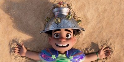 A smiling animated boy, Elio, lies on sandy ground with arms spread out. Wearing a colander hat with dangling light bulbs and a colorful, makeshift outfit, he looks cheerful and excited—ready for an Around the World Disney Parks adventure.