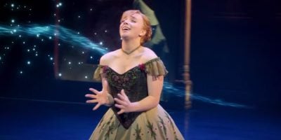 A woman in an ornate, off-shoulder dress stands on stage, singing passionately with her hands raised as blue magical sparkles swirl behind her—a scene reminiscent of Frozen: The Broadway Musical, now streaming on Disney+.