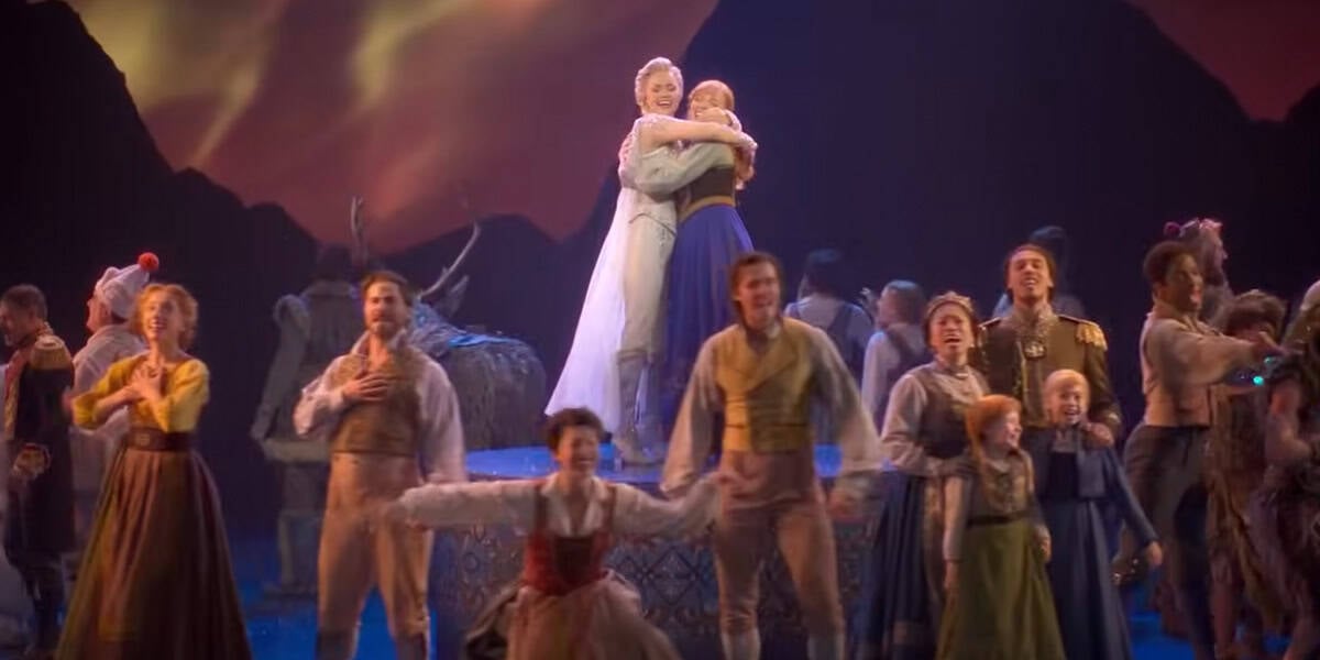A group of performers in colorful period costumes sing and dance on stage, with two women in the center embracing against a scenic mountain backdrop. The lively, joyful scene captures the magic of Frozen The Broadway Musical.