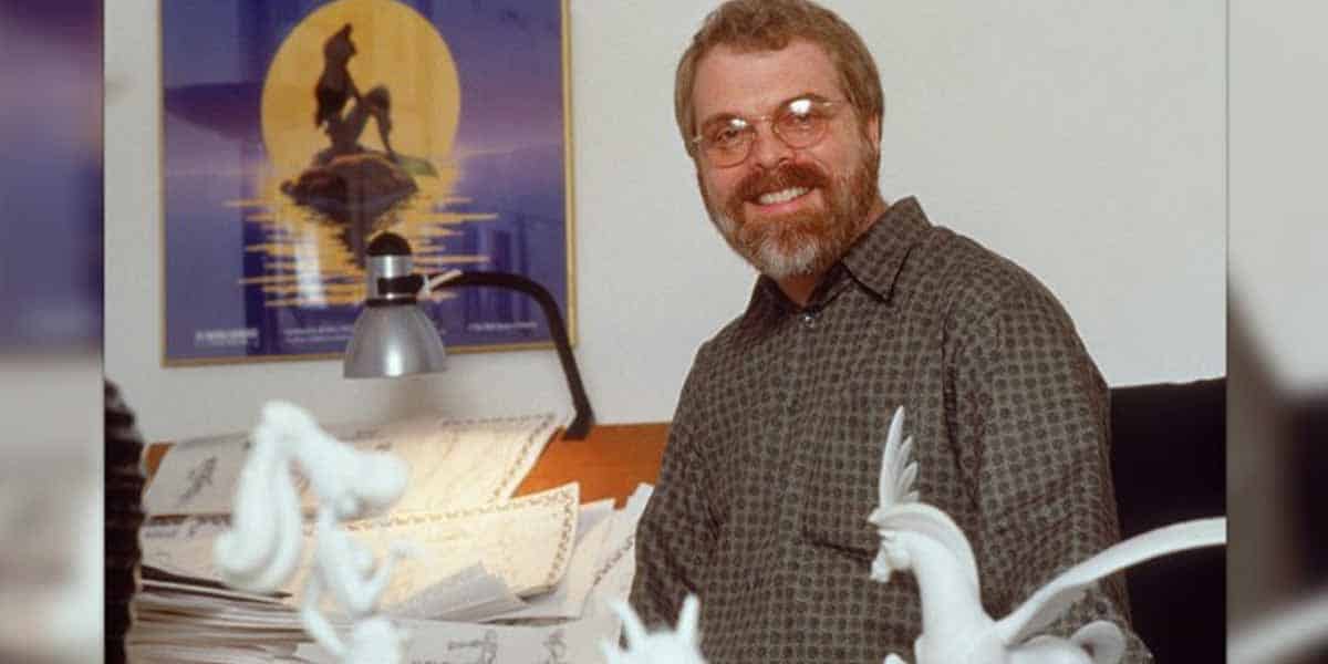 A man with glasses and a beard smiles while seated at a desk covered in papers and white sculpted figures, with a lamp and a framed poster featuring a character against a yellow moon—perhaps inspired by Ron Clements’ upcoming films—in the background.