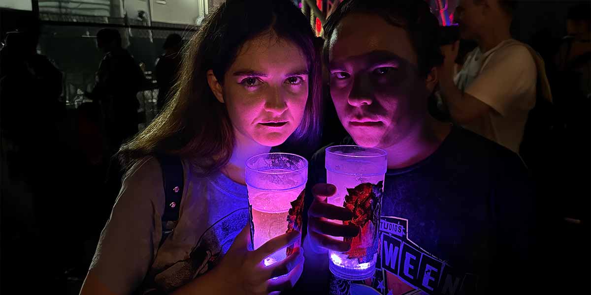 Two people stand close together at night at Halloween Horror Nights, holding glowing cups filled with light-up drinks. Their faces are illuminated by the purple and blue glow, with a crowd and dim lights visible in the background. Perfect for the easily spooked!.