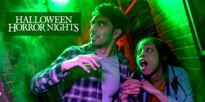 Two people who look easily spooked stand in a narrow, brick-walled hallway filled with eerie green light. “Halloween Horror Nights” appears in bold white text at the top left corner.