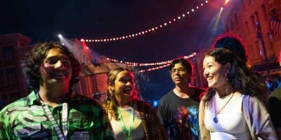 Four young people stand close together, smiling and talking under colorful string lights at night in an outdoor urban setting—like a beginner’s guide to fun before braving Halloween Horror Nights. The festive atmosphere buzzes with anticipation for the easily spooked at Universal Studios.