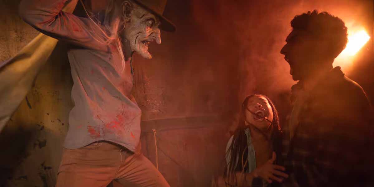 A scary figure with a distorted face and hat looms over two easily spooked people, one screaming in terror, in a dim, red-lit room with an eerie atmosphere—perfect for Halloween Horror Nights.