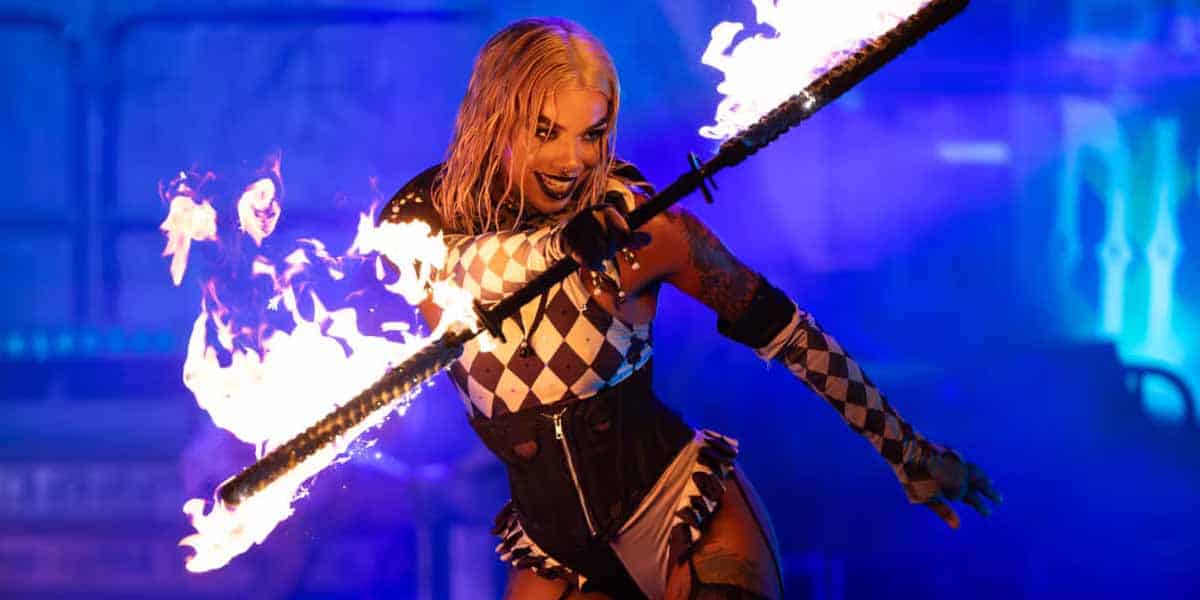 A performer in a black and white harlequin costume wields a flaming staff, smiling dramatically against a blue-lit backdrop—perfect for those who love Halloween Horror Nights but aren’t easily spooked.