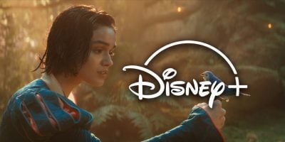A young woman in a blue dress gently holds a small blue bird on her hand in a lush forest, evoking the magic of the Snow White Remake. The Disney+ logo appears prominently over the image.