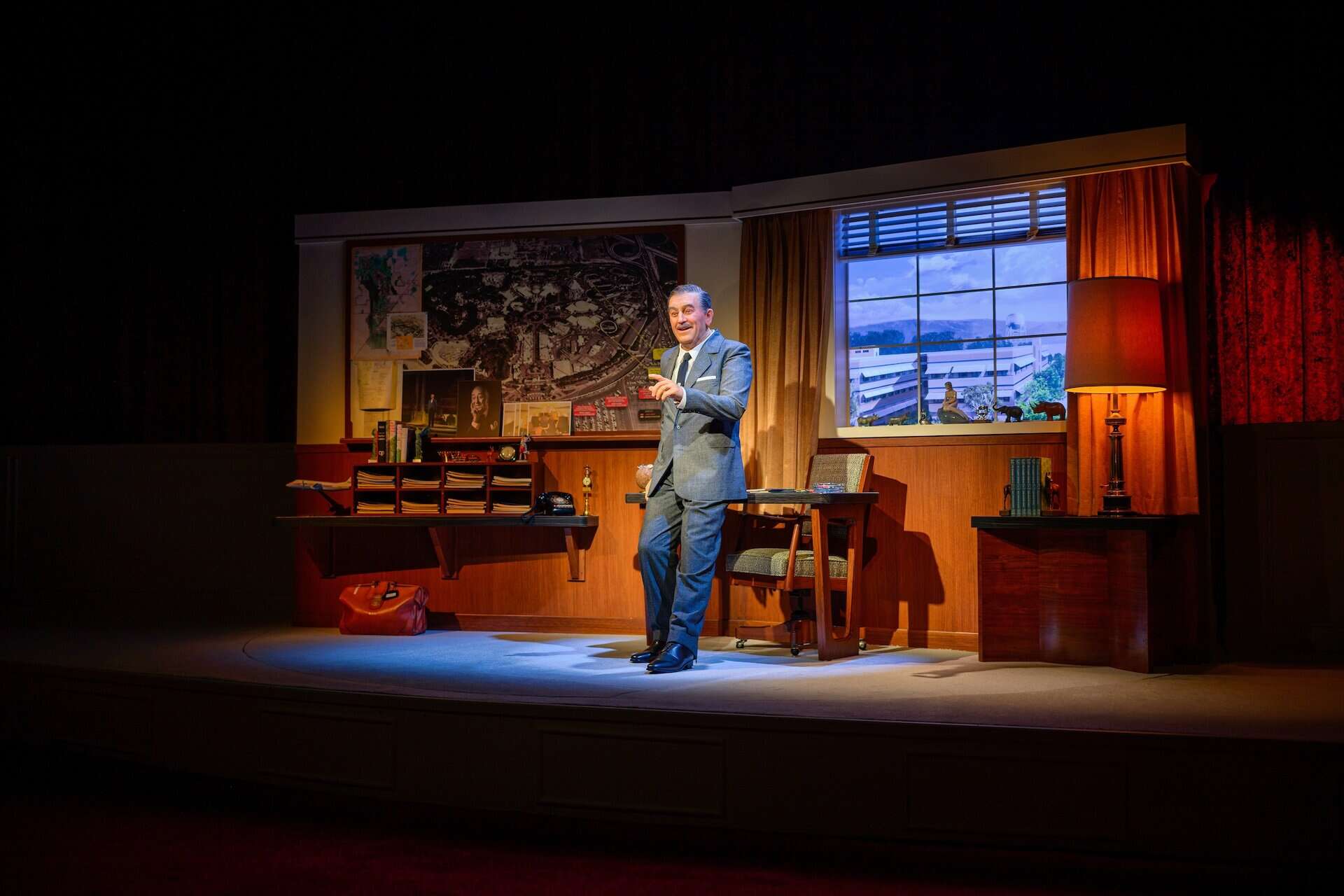 A man in a suit stands on a stage set designed as a mid-century office, with a desk, lamp, books, window, and various maps and photos—his stillness reminiscent of a Disney Animatronic—illuminated by a spotlight.