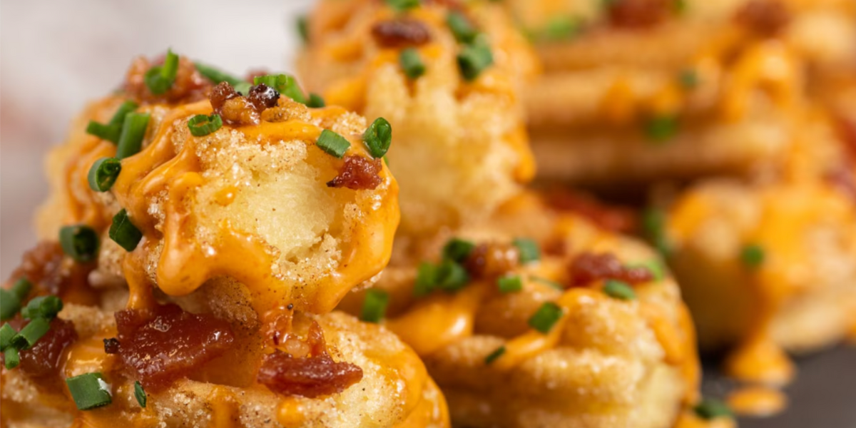 Close-up of crispy tater tots topped with melted cheddar cheese sauce, chopped chives, bits of bacon, and a side of sweet churros for the perfect savory and sweet snack combination.