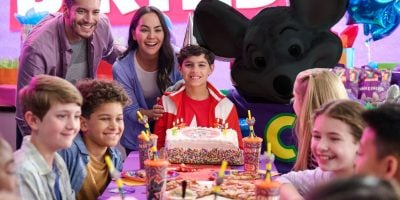 A group of kids sit around a birthday table with pizza, drinks, and a cake. Two adults and a mascot mouse in costume stand behind the smiling birthday child. Decor and balloons are in the background—no sign of credit card fraud here!.