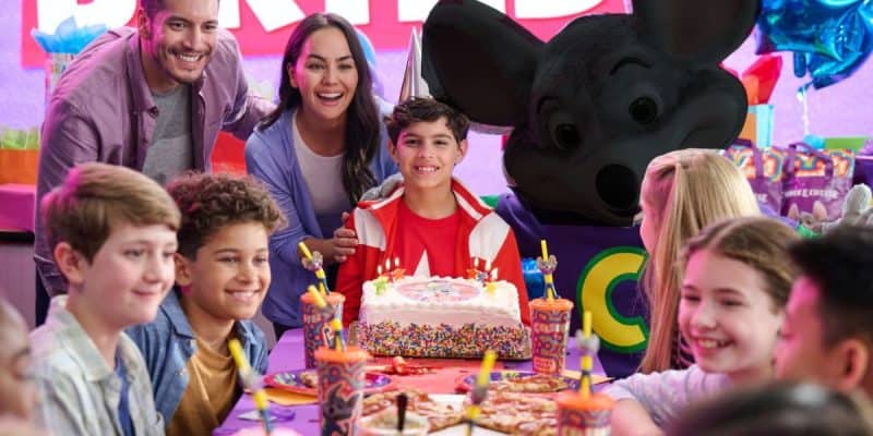 A group of kids sit around a birthday table with pizza, drinks, and a cake. Two adults and a mascot mouse in costume stand behind the smiling birthday child. Decor and balloons are in the background—no sign of credit card fraud here!.